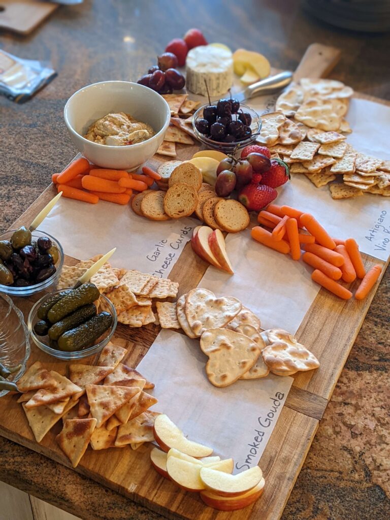 First steps in putting the charcuterie board together include adding crackers, fruit, veggies, and hummus.