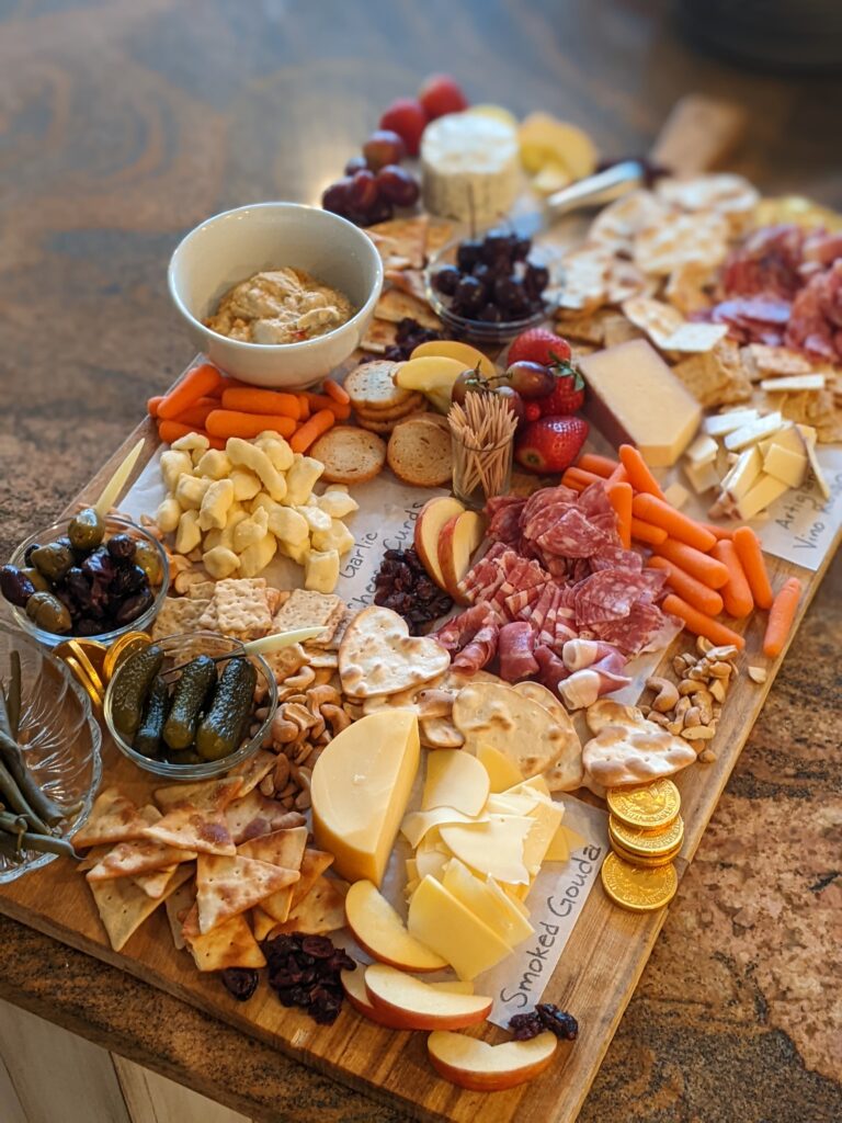 My completed charcuterie board with cheeses, fruits, veggies, pickles, olives, hummus, nuts, dried cranberries, sliced sopressata and prosciutto. 